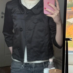 Svart kort kavaj med stora knappar - Snygg svart kavaj med bred krage och stora dekorativa knappar framtill. Jackan är kortare i modellen och har trekvartsärmar. Materialet är slätt och har en lätt glansig finish som ger en cool stil. Perfekt att styla med jeans eller kjol för en trendig look.