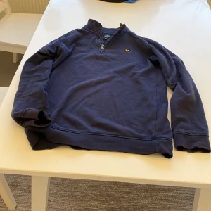 Marinblå Lyle & Scott half zip tröja - Snygg marinblå tröja från Lyle & Scott Junior med half zip och lång ärm. Tröjan har ett litet guldfärgat logomärke på bröstet och är tillverkad i mjuk bomull. Perfekt för dig som gillar klassisk stil med en sportig touch.