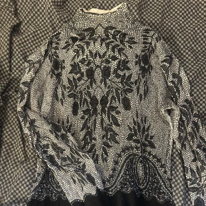 Svartvit blommig långärmad skjorta - Unik långärmad skjorta med svartvitt blommigt mönster och hög krage. Materialet känns mjukt och har en snygg struktur som sticker ut. Perfekt för dig som gillar detaljer och vill ha något som syns. Skjortan har en loose passform och coola kontraster.