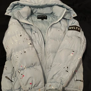Ljusblå pufferjacka från NVLTY - Säljer en ljusblå pufferjacka från NVLTY i storlek S. Jackan har coolt färgstänksmönster i svart, vitt och rosa. Den har en avtagbar huva. Perfekt för dig som vill sticka ut med en unik och trendig vinterjacka.