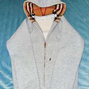 Grå Burberry hoodie med dragkedja - Snygg grå  Burberry hoddie med klassiskt rutigt foder i huvan. Tröjan har dragkedja framtill, snörning vid huvan och broderad logga på bröstet. Mjuk insida och ribbade muddar för extra komfort. Perfekt för dig som gillar stilrena och exklusiva plagg. PRIS KAN DISKUTERAS!