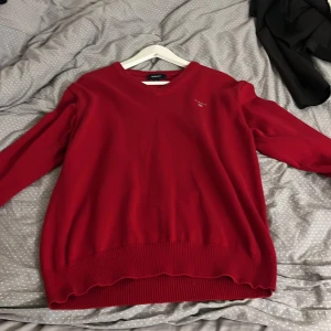 Röd stickad v-ringad tröja från GANT - Riktigt snygg röd stickad tröja från GANT med v-ringad hals och diskret logga broderad på bröstet. Tröjan har långa ärmar och ribbade muddar vid hals, ärmslut och nederkant. Perfekt för dig som gillar klassisk stil med en modern touch.