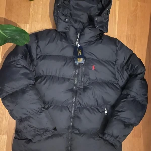 Svart pufferjacka  - Svart pufferjacka från Polo Ralph Lauren med huva och dragkedja framtill. Jackan har två sidofickor med dragkedja. Perfekt för kalla dagar med sin fluffiga och stilrena look.