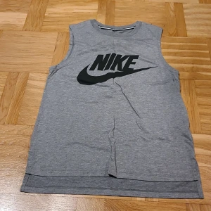 Grå ärmlös linnetopp från Nike XS - Säljer en grå ärmlös linnetopp från Nike i storlek XS. Toppen har en klassisk rund hals och stort svart Nike-logotryck på framsidan. Tillverkad i mjuk bomull, perfekt för träning eller chill. Enkel och sportig design som funkar året runt.