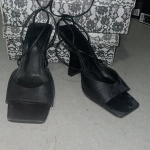 Svarta klackade sandaler med snörning - Svarta sandaler från H&M med fyrkantig tå, bred rem över foten och höga blockklackar. Skorna har tunna snören som lindas runt vristen för en trendig look. Tillverkade i syntetmaterial med stilren design som passar perfekt till sommarens outfits.