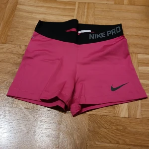 Nike Pro rosa träningsshorts S - Nike Pro shorts i stark rosa med svart bred midja och logga. Tight passform, korta ben och elastiskt material som andas. Perfekta för gymmet eller sport, med Dri-FIT-teknologi för att hålla dig torr. Snyggt Nike Pro-tryck på midjan.