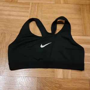Svart sporttopp från Nike - Svart sporttopp från Nike med klassisk vit logga framtill. Modellen är ärmlös med brottarrygg och bred resår nedtill för extra stöd. Materialet är mjukt och stretchigt polyester, perfekt för träning och aktiva dagar.