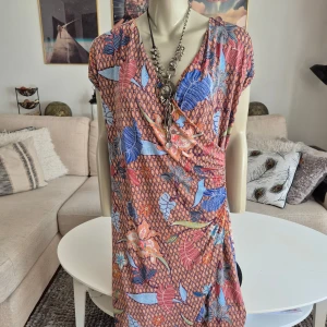 Pearl * Flerfärgad klänning * stl L - XL - Färgglad klänning med ett vackert blommönster i blått, rött och orange. * Omlottdesign framtill och korta ärmar. * Stl L men passar också XL. Stretchig Se mått nedan. * Nyskick. * Mått: - Längd ca: 100 cm - Armhåla till armhåla ca: 55 cm  Djur och rökfritt hem. Se bilder och förstora då de ingår i beskrivningen. Se gärna mina övriga annonser.   Vid avhämtning finns den i Sollentuna.