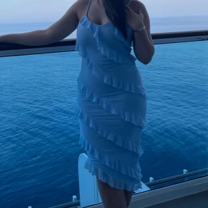 Ljusblå volangklänning med axelband - Säljer en ljusblå klänning med smala axelband och flera volangdetaljer som går diagonalt över hela klänningen. Modellen är figurnära och har en snygg, feminin siluett som passar perfekt för sommarkvällar. Klänningen slutar strax under knäna och har en fräsch vibe.