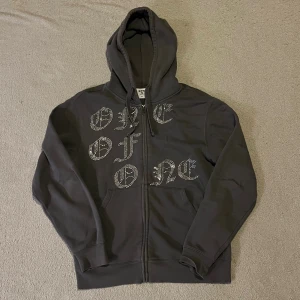 Grå One of One kofta - Grå kofta från One of One med glittrande paljetter. Tröjan har huva, lång ärm och praktisk dragkedja framtill. Perfekt för dig som gillar streetwear och vill sticka ut med coola detaljer.