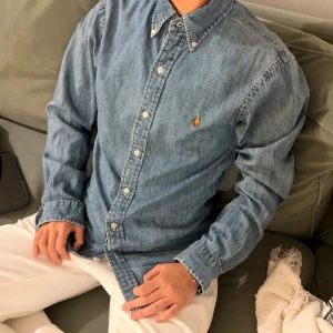 Blå jeansskjorta från Polo Ralph Lauren - Snygg blå jeansskjorta från Polo Ralph Lauren i slim fit. Skjortan är tillverkad i 100% bomull och har klassisk krage med knappar, samt den ikoniska broderade loggan på bröstet. Perfekt för dig som gillar en clean och tidlös look.