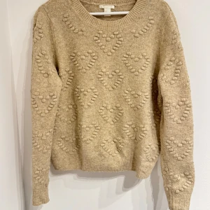 Beige stickad tröja med hjärtmönster H&M - Supermysig beige stickad tröja från H&M med upphöjt hjärtmönster över hela tröjan. Rund halsringning, ribbade muddar och långärmad modell. Perfekt för dig som gillar gulliga detaljer och vill ha en varm tröja till hösten och vintern.