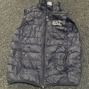 Svart dunväst från EA7 Emporio Armani - Snygg svart dunväst från EA7 Emporio Armani med quiltad design och hög krage. Västen har dragkedja framtill med logga på zip-pullern och broderad logga på bröstet. Perfekt för lager-på-lager och streetstyle.