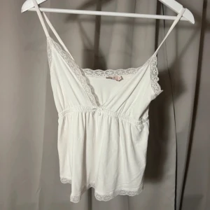 Vit spetskantad linnetopp från Nelly - Supersöt vit linnetopp från Nelly i storlek XS. Toppen har tunna axelband, v-ringning och är dekorerad med spetsdetaljer längs halsringning och nederkant. Mjuk och lätt bomullskänsla, perfekt för varma dagar. Snyggt rynkad under bysten för en feminin siluett. Buda gärna❤️är ni inte trevliga så tillåter jag tyvärr inte köp🫶OBS! Har sytt upp banden på den!! 