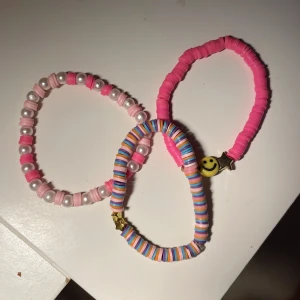 Färgglada pärlarmband med smiley - Tre elastiska armband med pärlor i rosa, vitt och pastellfärger. Ett armband har vita pärlor och rosa skivor, ett är helrosa med en gul smiley och stjärna, och det tredje har randiga pastellfärgade skivor och små guldstjärnor. Perfekt för att liva upp din stil!