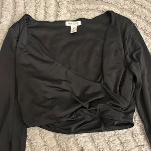 Svart omlottopp från NLY ONE - Säljer en svart omlottopp från NLY ONE i mjukt och glansigt material. Toppen har lång ärm, croppad passform och snygg omlottdetalj framtill. Perfekt för dig som gillar stilrena och trendiga plagg med en twist.