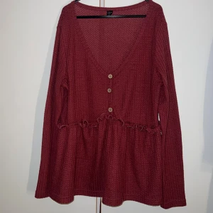 Vinröd blus med volang från Shein Curve - Säljer en vinröd blus från Shein Curve i storlek XL. Blusen har v-ringning, tre träknappar framtill och en söt volangdetalj vid midjan. Materialet är mjukt och strukturerat, med lång ärm och loose passform. Perfekt för dig som gillar bekväma och snygga toppar.