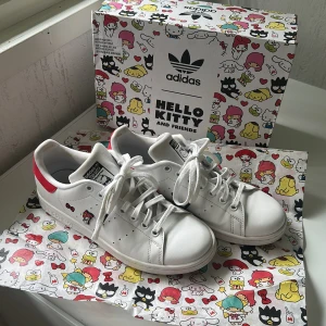 Adidas Stan Smith Hello Kitty sneakers - (SKICKAS NYTVÄTTADE) Unika Adidas Stan Smith Hello Kitty & Friends sneakers i vitt skinn med röda detaljer och Hello Kitty-figurer på sidorna. Klassisk rund tå, platt sula och snörning. Kommer med originalkartong och specialdesignat papper med Hello Kitty-motiv. Perfekt för dig som älskar limited edition och kawaii-stil.