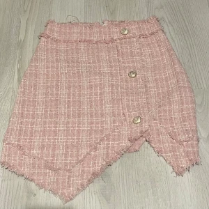 Rutig rosa kortkjol med knappar - Säljer en rosa kortkjol med rutigt mönster och råa kanter. Kjolen har en unik asymmetrisk nederkant och tre dekorativa guldfärgade knappar framtill. Materialet känns som en blandning av bomull och polyester, perfekt för vår och sommar.