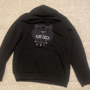 Svart Kenzo hoodie i bra kondition  - Svart hoodie från Kenzo med ikoniskt tigerbroderi och Kenzo Paris-text på bröstet. Tröjan har huva, magficka och långa ärmar. Materialet är mjukt och känns som bomull, perfekt för chill dagar. Snygg och stilren design som sticker ut med detaljerad brodyr.