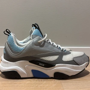 Dior b22  - Chunky Dior sneakers med vit, grå och blå färg. Ovandel i mesh och skinn med coolt mönstrade detaljer och snörning. Sulan är tjock och har inslag av svart och blått, vilket ger en sportig vibe. Perfekta för dig som gillar statement-skor med lyxig känsla.