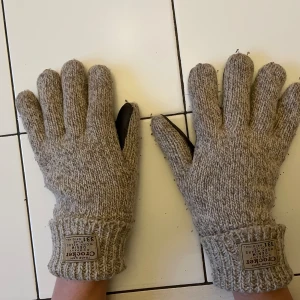 Stickade handskar med skinn från Crooked - Snygga stickade handskar från Crooked med beige yta och mörkbruna skinninsidor i handflatan. De har ribbstickade muddar och en patch med logga på ovansidan. Perfekta för kalla dagar när du vill ha både stil och funktion.