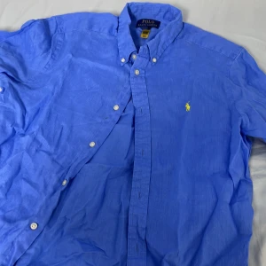 Blå linneskjorta från Polo Ralph Lauren passar S-M - Snygg blå skjorta från Polo Ralph Lauren i linne med klassisk button down-krage och vita knappar. Ikoniska gula broderade loggan på bröstet. Perfekt för dig som gillar stilren och avslappnad look.