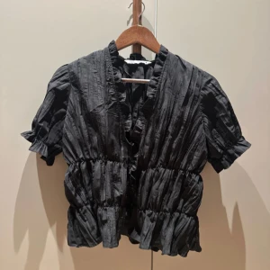 Svart blus med volang från Zara - Svart blus från Zara med korta puffärmar och volangdetaljer längs krage och nederkant. Blusen har en lätt glansig yta och struktur i tyget, samt markerad midja för en feminin siluett. Perfekt att styla med jeans eller kjol.