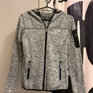 Grå fleecetröja med huva från OTP - Gråmelerad fleecetröja från OTP med huva och svarta detaljer. Jackan har hel dragkedja framtill, två fickor med dragkedja och en extra ficka med dragkedja på ärmen. Perfekt för chill dagar eller när du vill vara bekväm men ändå se fräsch ut. Köpte från Stadium i storlek s