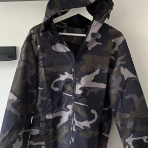Camouflage windbreaker  från Lindfors - Snygg regnjacka från Lindfors med camouflagemönster i mörka toner. Jackan har huva, dragkedja framtill och elastiska muddar vid ärmsluten. Perfekt för regniga dagar och streetstyle-vibe. Materialet är lätt och vattenavvisande syntet.
