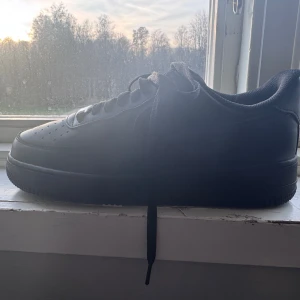 Svarta Nike Air Force 1 sneakers - Säljer ett par svarta Nike Air Force 1 sneakers med klassisk låg siluett. Skorna har snörning, perforerad tå för ventilation och en platt sula. Tillverkade i skinn med diskret Nike-logga på sidan och metallbricka med 'AF1' på snörningen. Perfekt för dig som gillar streetwear. Nypris: 1399kr