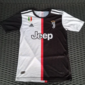 Juventus Ronaldo 7 fotbollströja Adidas - Juventus matchtröja med Ronaldo 7 på ryggen, tillverkad i lätt och ventilerande Climalite-material. Snygg design med svart och vit färgblock, röd vertikal linje, broderat Juventus-emblem med tre guldstjärnor och Jeep-logga på bröstet. Klassiska Adidas-ränder på axlarna.
