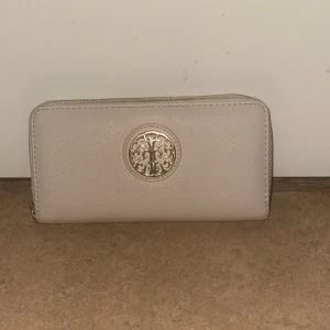 Beige plånbok med dragkedja - Snygg beige plånbok med silverfärgat logomärke på framsidan. Plånboken har dragkedja runtom och flera fack för kort och mynt. Materialet är syntet med lätt glansig finish och insidan är svart. Perfekt storlek för att ha med sig överallt.