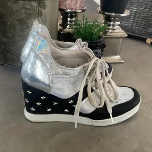  Cooolaaaa Silvriga sneakers med stjärnor och klack - Unika sneakers med silvriga detaljer, vit mesh och svart parti med silverfärgade stjärnor på kilklacken. Skorna har snörning och holografisk häl. Perfekt för dig som gillar att sticka ut med coola detaljer och höjd.
