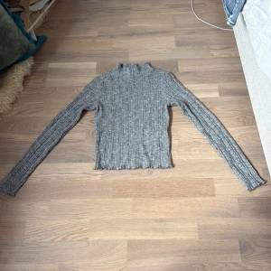 Grå ribbad från FB Sister - Gråmelerad ribbad från FB Sister i storlek XXS. Tröjan har lång ärm, hög hals och är stickad i mjukt material. Perfekt för lager-på-lager och har en slim passform som sitter snyggt. Enkel att matcha med jeans eller kjol.