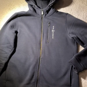 Svart hoodie från Sail Racing (Använd 3 gånger) - Mörkgrå hoodiejacka från Sail Racing med dragkedja framtill och bröstficka med dragkedja. Jackan har en mjuk fleeceinsida, huva med broderad logga och stilren design. Perfekt för kyliga dagar och enkel att matcha med det mesta.