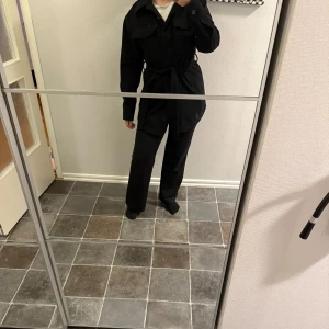 Svart kapppa med bälte - Säljer en svart overshirt med bälte i midjan och stora bröstfickor. Jackan har klassisk krage, knappar framtill och långa ärmar med knapp vid ärmslut. Perfekt jacka nu till kyliga höst dagar! Jackan är flitigt använd men i mycket bra skick överlag.
