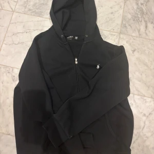 Svart hoodie från Polo Ralph Lauren - Svart hoodie från Polo Ralph Lauren med dragkedja och klassisk vit logga på bröstet. Tröjan har huva, känguruficka och är tillverkad i mjuk bomull. Perfekt för chill dagar eller när du vill ha en clean streetstyle-look.