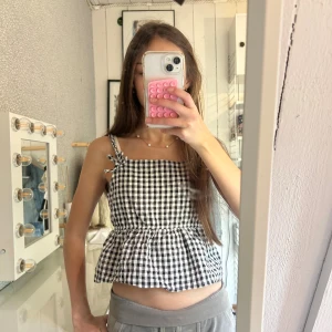Rutig blus med volang från H&M - Säljer den här gulliga toppen från H&M för 50 kr. Svartvit rutigt mönster, smock i ryggen och volang nertill som gör att den sitter fint. Smala axelband som knyts i rosetter.  Storlek 152 (11–12 år). Jag är 158 cm och den passar perfekt. Använd väldigt lite, i jättebra skick.