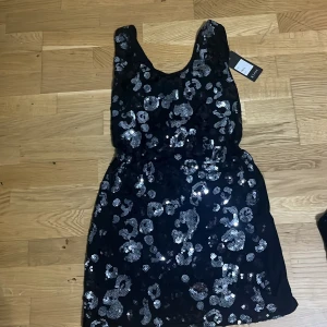 Svart paljettklänning med rosetter KIAH - Svart kort klänning från KIAH med glittriga silverpaljetter och två stora rosetter på ryggen. Klänningen har ärmlös design, rund halsringning och en snygg cut-out detalj i ryggen. Perfekt för dig som vill sticka ut med en unik och festlig look, prislappen e kvar på klänningen  