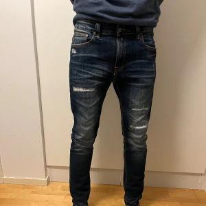 Nudie jeans - Säljer ett par mörkblå jeans från Nudie med slitna detaljer och snygga kontrastsömmar. Jeansen har klassiska fem fickor och Nudie-logga på bakfickorna. Storlek 34/32