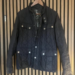 Mörkblå quiltad jacka från Barbour - Quiltad mörkblå jacka från Barbour International med snygga silverfärgade knappar och flera fickor framtill. Jackan har ståkrage, dragkedja och tryckknappar. Perfekt för dig som gillar klassisk stil med modern touch.