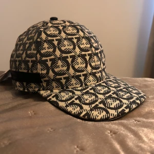 Salvatore Ferragamo beige keps - Snygg keps från Salvatore Ferragamo i beige och svart med ikoniskt mönster över hela kepsen. Justerbar rem baktill med metallspänne och läderdetalj. Tillverkad i Italien och gjord i bomullsmaterial. Perfekt för dig som vill sticka ut med en lyxig accessoar.