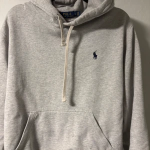 Grå hoodie från Polo Ralph Lauren - Snygg ljusgrå hoodie från Polo Ralph Lauren med klassisk broderad logga på bröstet. Tröjan har huva med snörning, känguruficka och half zip-dragkedja. Perfekt för chill dagar eller när du vill ha en clean och stilren look.