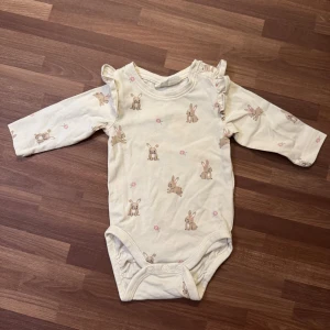Body med kaniner - Supersöt krämvit body från Kappahl i storlek 56 (1-2mån). Använd en gång, utan anmärkningar. Långärmad modell med volangdetaljer på axlarna och praktiska tryckknappar vid axel och gren.
