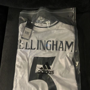 Vit Real Madrid Bellingham tröja Adidas - Snygg vit Real Madrid fotbollströja med Bellingham och nummer 5 på ryggen. Tröjan har svart tryck och klassisk rund hals med svart/vit detalj. Tillverkad i polyester och perfekt för dig som vill bära ditt favoritlag med stil.