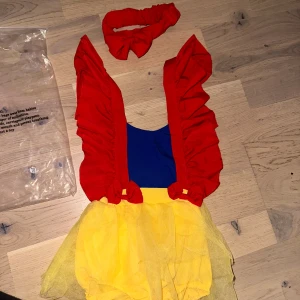 Snövit-inspirerad babyklänning  body med rosett med tillhörande röd pannband - Söt Snövit-inspirerad babyklänning med matchande hårband. Klänningen har en blå överdel, röda puffärmar och en gul kjol med tyll. Perfekt för kalas eller maskerad! Supersöt babyklänning inspirerad av Snövit med blå överdel, röda volangärmar och rosetter samt gul tyllkjol. Matchande röd hårband med rosett ingår. Perfekt för temadagar eller maskerad! 🎁 