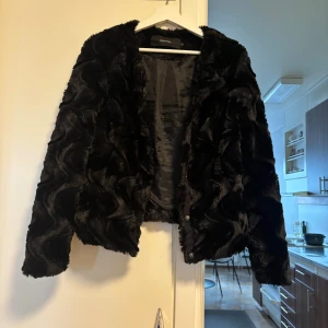 Svart pälsjacka från Vero Moda - Säljer en svart pälsjacka från Vero Moda med snyggt vågigt mönster i pälsen. Jackan har långärmade ärmar och är fodrad på insidan för extra komfort. Perfekt för dig som vill ha en cool och trendig look under höst och vinter.
