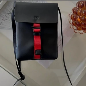 Svart axelväska från Gaston Luga - NY! - Minimalistisk svart axelväska från Gaston Luga med röd rem framtill och justerbar axelrem. Väskan har stilren design med lock och präglad logga på framsidan. Materialet är slätt syntet och väskan har en kompakt, rektangulär form som passar till det mesta.