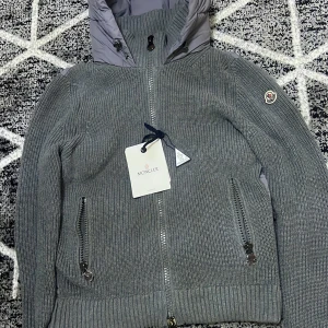 Grå puffer moncler cardigan  - Snygg grå pufferjacka från Moncler med stickade ärmar och ribbade muddar. Jackan har quiltad baksida och huva i glansigt material, dragkedjor framtill och på fickorna samt Moncler-logga på ärmen. Perfekt för kyliga dagar med en modern vibe.